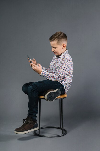 Ein Junge, der auf Schemel unter Verwendung des Handys gegen grauen Hintergrund sitzt