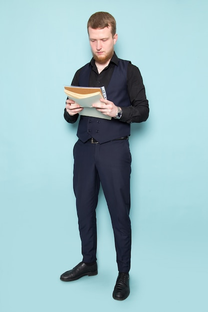 Ein junger attraktiver Mann der Vorderansicht mit Bart im schwarzen dunklen klassischen modernen Anzug, der Akten auf dem blauen Raum hält
