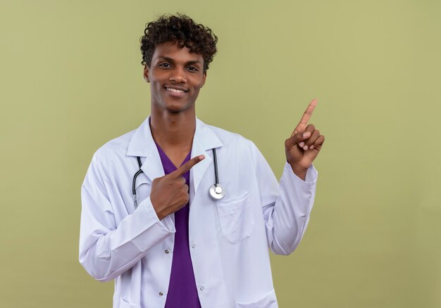Ein junger aufgeregter gutaussehender dunkelhäutiger männlicher Arzt mit lockigem Haar, der weißen Mantel mit Stethoskop trägt, das mit Zeigefingern auf einer Grünfläche nach oben zeigt