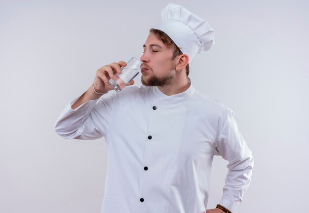 Ein junger bärtiger Kochmann, der weiße Kochuniform und Hut trägt, trinkt ein Glas Wasser, während er auf eine weiße Wand schaut