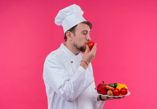 Ein junger bärtiger Kochmann in der weißen Uniform, die einen Teller mit frischem Gemüse hält, während Tomate auf einer rosa Wand riecht