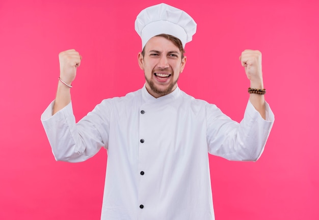 Ein junger bärtiger Kochmann in der weißen Uniform, die geballte Fäuste in der Luft erhebt, während er auf eine rosa Wand schaut