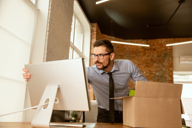 Ein junger Geschäftsmann, der im Büro umzieht und einen neuen Arbeitsplatz bekommt. Junger kaukasischer männlicher Büroangestellter rüstet neues Kabinett nach Beförderung aus. Sieht Glücklich aus. Geschäft, Lebensstil, neues Lebenskonzept.