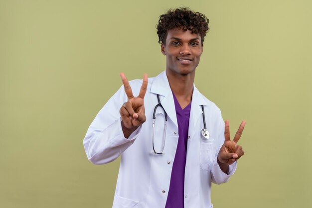 Ein junger gutaussehender dunkelhäutiger Mann mit lockigem Haar, der einen weißen Kittel mit Stethoskop trägt, der zwei Finger zeigt und auf einer Grünfläche lächelt