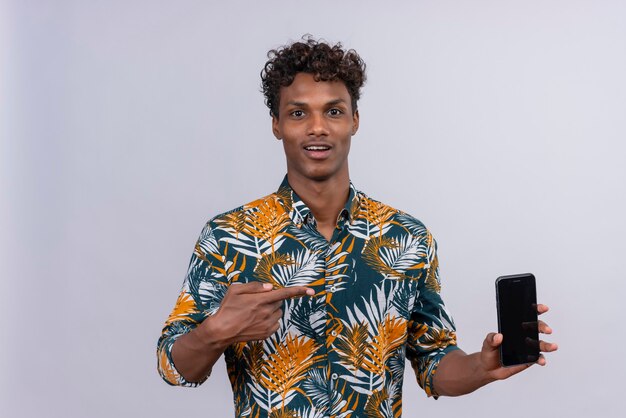 Ein junger gutaussehender dunkelhäutiger Mann mit lockigem Haar im Blatt bedruckte Hemd, das auf Mobiltelefon mit Zeigefinger zeigt