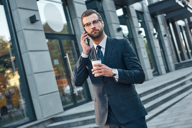 Ein junger gutaussehender geschäftsmann mit brille telefoniert und trinkt kaffee