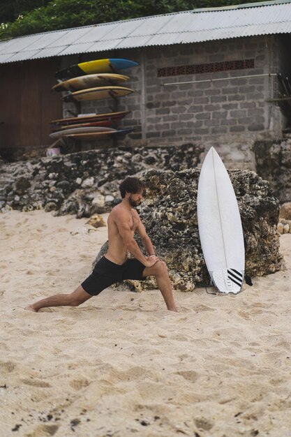 Ein junger gutaussehender Surfer am Meeresufer wärmt sich vor dem Surfen auf. Übungen vor dem Sport, Stretching vor dem Surfen.