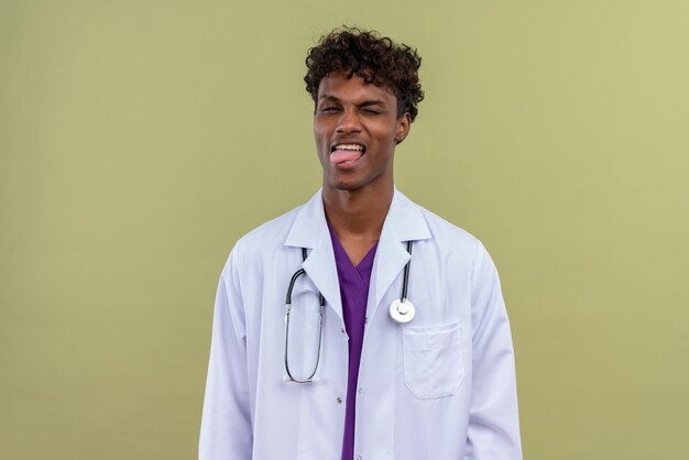 Ein junger hübscher dunkelhäutiger Arzt mit lockigem Haar, der einen weißen Kittel mit Stethoskop trägt und seine Zunge auf einer Grünfläche zeigt
