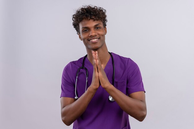 Ein junger hübscher dunkelhäutiger Arzt mit lockigem Haar in violetter Uniform und Stethoskop, das in der dankbaren Geste die Hände zusammenhält