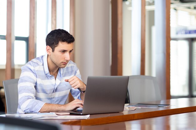 Ein junger hübscher Mann der Vorderansicht im gestreiften Hemd, der innerhalb des Konferenzsaals unter Verwendung seines silbernen Laptops während des Tagesarbeitsaktivitätsgebäudes arbeitet