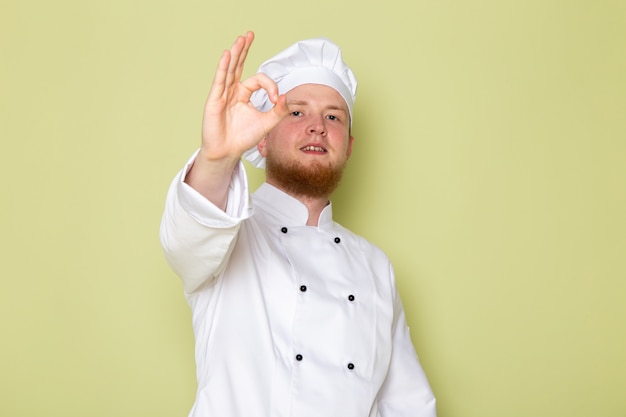 Ein junger männlicher Koch der Vorderansicht in der weißen Kopfkappe des weißen Kochanzugs, der gutes Zeichen zeigt