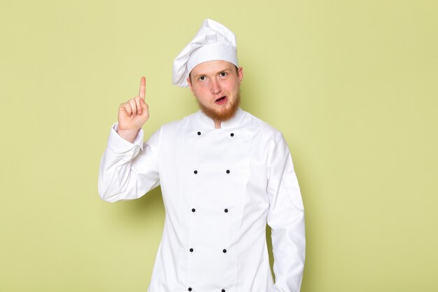 Ein junger männlicher Koch der Vorderansicht in der weißen Kopfkappe des weißen Kochanzugs mit erhöhtem Finger