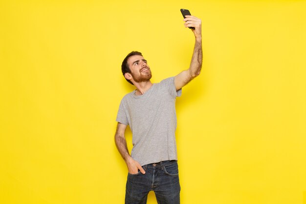 Ein junger Mann der Vorderansicht im grauen T-Shirt, das ein Selfie auf der gelben Wandmann-Farbmodell-Emotionskleidung nimmt