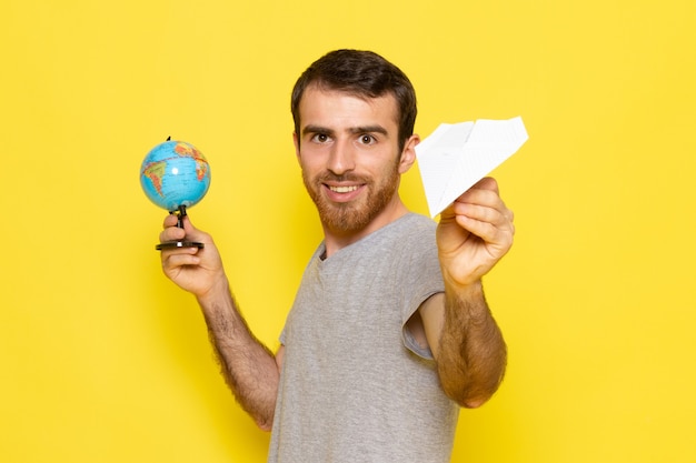 Ein junger Mann der Vorderansicht im grauen T-Shirt, das kleinen Globus und Papierflugzeug auf dem gelben Schreibtischmann-Farbmodell hält