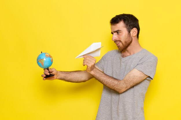 Ein junger Mann der Vorderansicht im grauen T-Shirt, das kleinen Globus und Papierflugzeug auf dem gelben Wandmann-Farbmodell hält