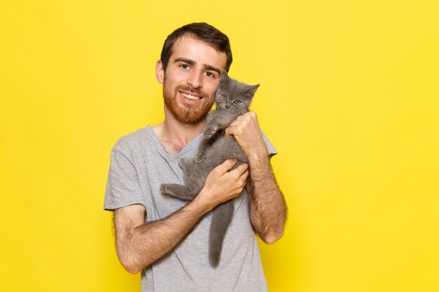 Ein junger Mann der Vorderansicht im grauen T-Shirt, das niedliches graues Kätzchen auf dem gelben Wandmannausdruck-Emotionsfarbmodell hält