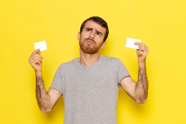Ein junger Mann der Vorderansicht im grauen T-Shirt, das weiße Karten auf dem gelben Wandmannausdruck-Emotionsfarbmodell hält