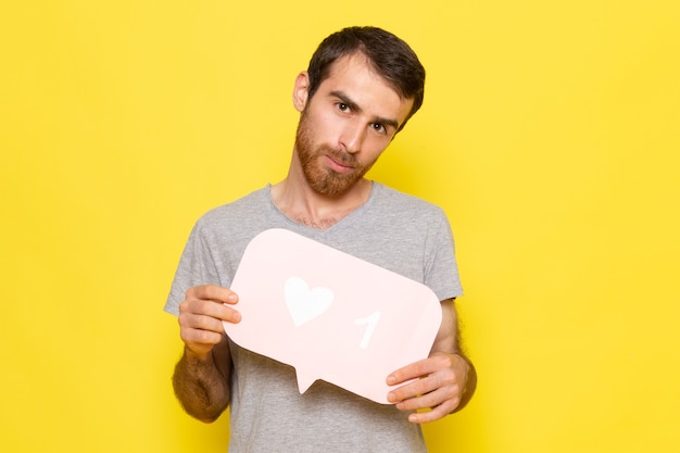 Ein junger Mann der Vorderansicht im grauen T-Shirt, das weißes Zeichen auf der gelben Wandmannausdruck-Emotionsfarbe hält