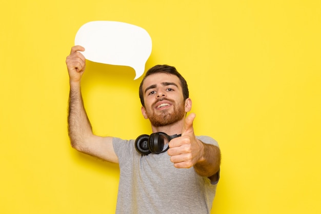 Ein junger Mann der Vorderansicht im grauen T-Shirt, das weißes Zeichen mit Lächeln auf dem gelben Wandmannausdruck-Emotionsfarbmodell hält