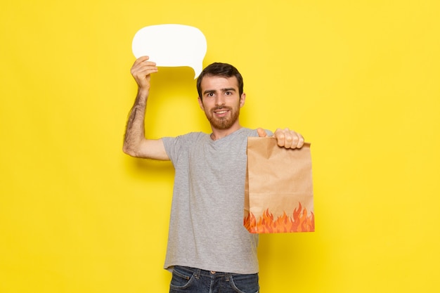 Ein junger Mann der Vorderansicht im grauen T-Shirt, das weißes Zeichen und Paket mit Lächeln auf dem gelben Wandmannausdruck-Emotionsfarbmodell hält