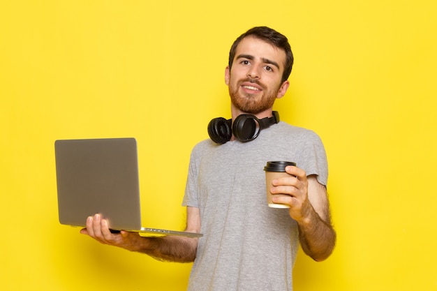 Ein junger Mann der Vorderansicht im grauen T-Shirt, der Kaffeetasse hält und Laptop mit Lächeln auf dem gelben Wandmannausdruck-Emotionsfarbmodell verwendet