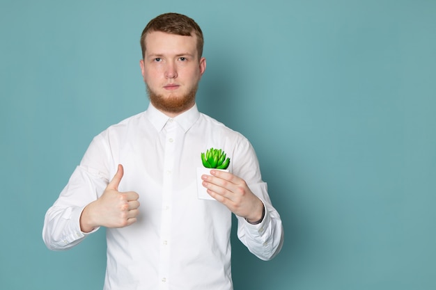 Ein junger Mann der Vorderansicht im weißen Hemd, das kleine grüne Pflanze auf dem blauen Raum hält