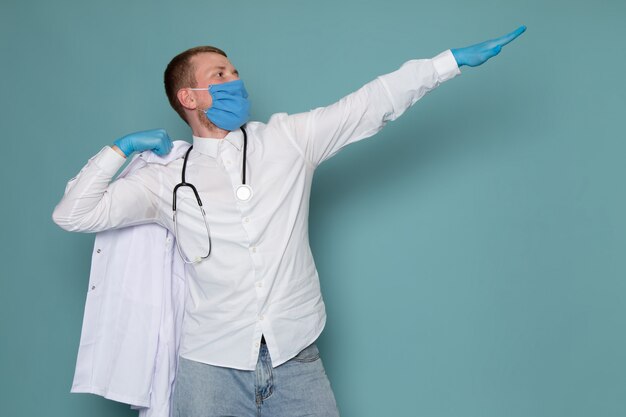 Ein junger Mann der Vorderansicht im weißen Hemd stethoskopiert blaue Handschuhe und Maske, die auf dem blauen Schreibtisch aufwirft