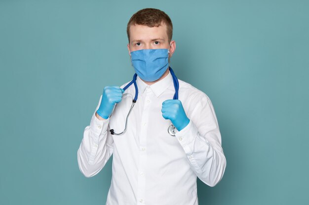 Ein junger Mann der Vorderansicht im weißen Hemd und in den blauen Handschuhen mit blauer Maske auf dem blauen Raum