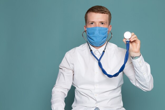 Ein junger Mann der Vorderansicht im weißen Hemd und in der blauen Maske mit Stethoskop auf dem blauen Raum