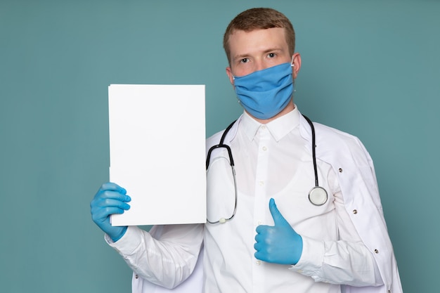 Ein junger Mann der Vorderansicht im weißen medizinischen Anzug blaue Handschuhe und Maske auf dem blauen Schreibtisch
