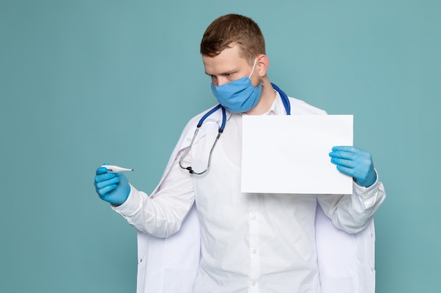 Ein junger Mann der Vorderansicht im weißen medizinischen Anzug mit Handschuhen und Maske auf dem blauen Schreibtisch