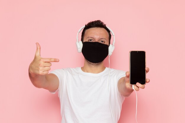 Ein junger Mann der Vorderansicht in der schwarzen Maske des weißen T-Shirts, die Musik durch Kopfhörer auf der Farbemotionshaltung des rosa Schreibtischmanns hört