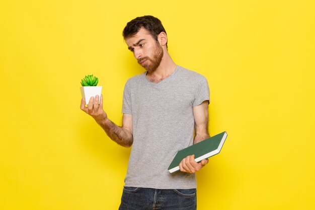 Ein junger Mann der Vorderansicht in grauem T-Shirt, das Heft und Pflanze auf dem gelben Wandmannausdruck-Emotionsfarbmodell hält