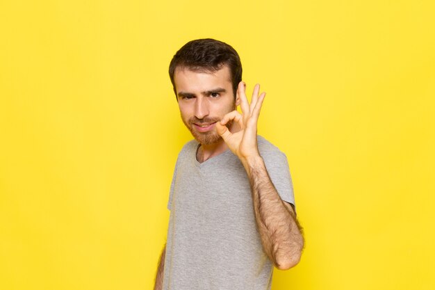 Ein junger Mann der Vorderansicht in grauem T-Shirt, das in Ordnung Zeichen auf der gelben Wandmann-Farbmodell-Emotionskleidung zeigt