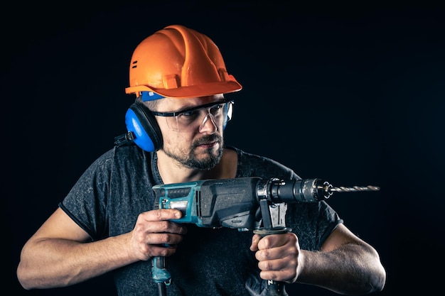 Ein junger Mann im Schutzhelm des Overalls hält eine elektrische Bohrmaschine