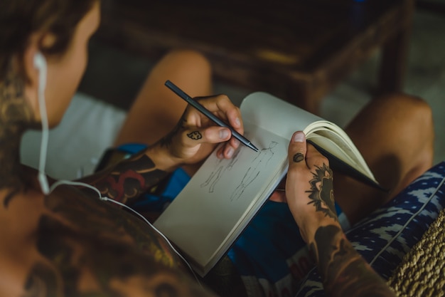 Ein junger Mann in Tattoos mit Kopfhörern hört Musik und zeichnet ein Notizbuch.