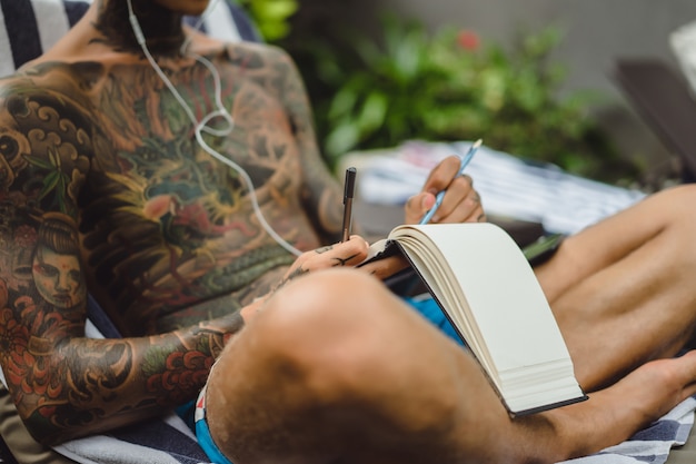 Ein junger Mann in Tattoos mit Kopfhörern hört Musik und zeichnet ein Notizbuch.