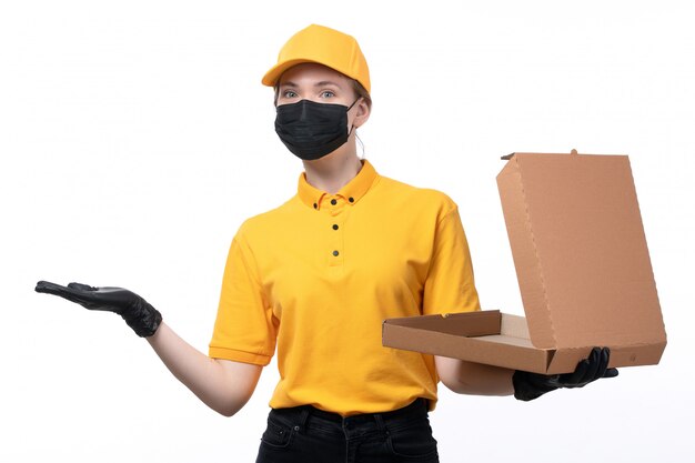 Ein junger weiblicher Kurier der Vorderansicht in den gelben einheitlichen schwarzen Handschuhen und in der schwarzen Maske, die eine leere Pizzaschachtel halten