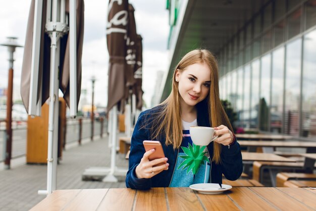 Ein junges hübsches Mädchen mit langen Haaren sitzt draußen im Café am Tisch. Sie trägt eine blaue Jacke. Sie hält eine Tasse Kaffee und lächelt in die Kamera.