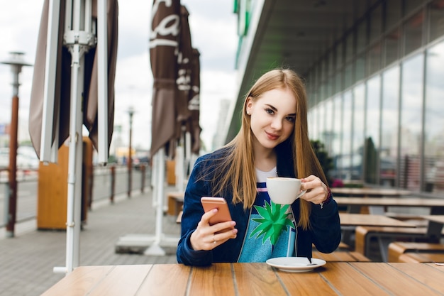 Ein junges hübsches Mädchen mit langen Haaren sitzt draußen im Café am Tisch. Sie trägt eine blaue Jacke. Sie hält eine Tasse Kaffee und lächelt in die Kamera.