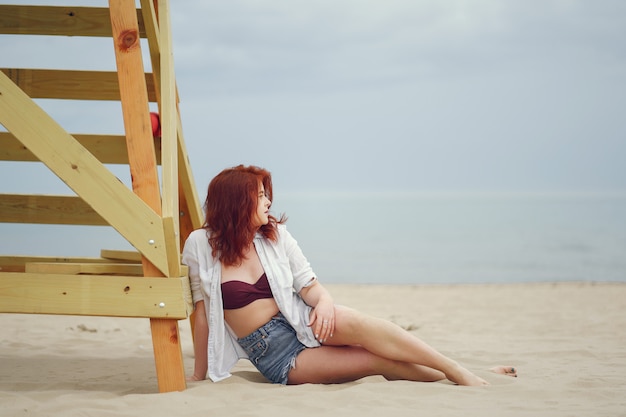 Ein junges redheaded Mädchen in einem weißen Hemd und in den kurzen Jeanshosen, die auf dem Sand auf dem Strand sitzen