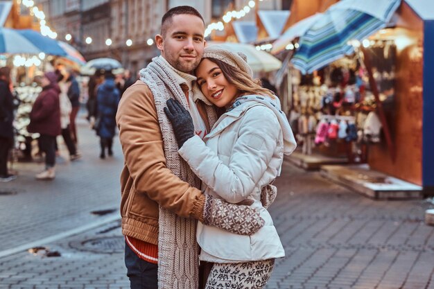 Ein junges romantisches Paar, das warme Kleidung trägt und sich zur Weihnachtszeit in der Abendstraße im Freien umarmt