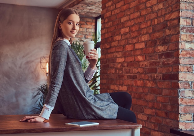 Ein junges schlankes sexy Mädchen in einem grauen Kleid hält eine Tasse Kaffee sitzen