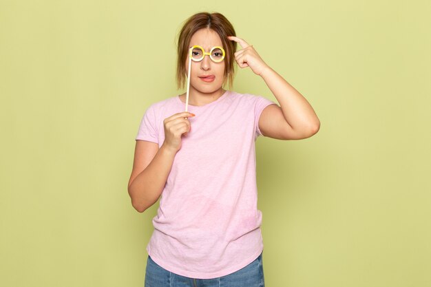 Ein junges schönes Mädchen der Vorderansicht in den blauen Jeans des rosa T-Shirts, das mit der Sonnenbrille des Spielzeugsticks auf Grün aufwirft