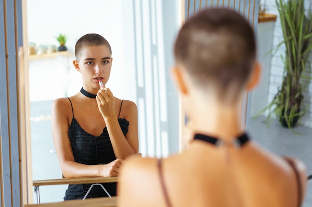 Ein junges schönes Mädchen mit kurzen Haaren malt ihre Lippen und betrachtet das Spiegelbild in schwarzen Kleidern