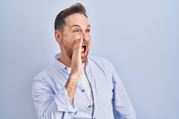 Ein kaukasischer Mann mittleren Alters steht über einem blauen Hintergrund und schreit und schreit laut von Seite zu Seite mit der Hand auf den Mund Kommunikationskonzept