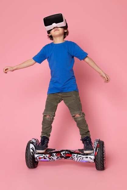 Ein kleiner Junge der Vorderansicht im blauen T-Shirt, das vr auf Segway auf dem rosa Raum spielt