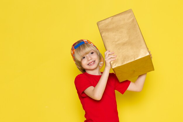 Ein kleiner Junge der Vorderansicht in der bunten Kappe des roten T-Shirts und der grauen Jeans, die Box auf dem gelben Hintergrund halten