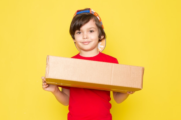 Ein kleiner Junge der Vorderansicht in der bunten Kappe des roten T-Shirts und in der Khakihose, die Box auf dem gelben Hintergrund hält