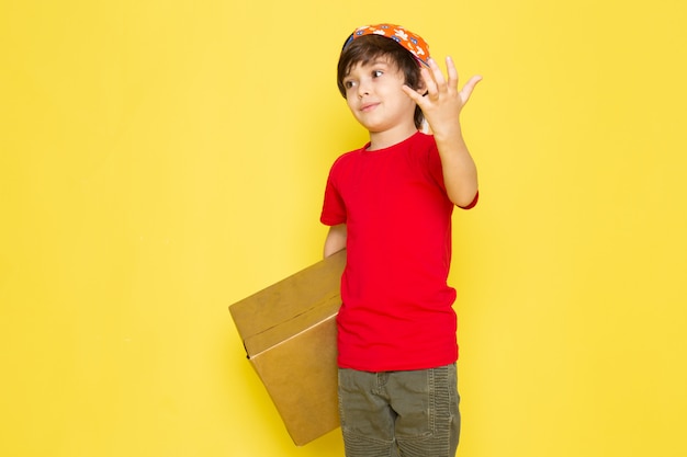 Ein kleiner Junge der Vorderansicht in der bunten Kappe des roten T-Shirts und in der Khakihose, die Box auf dem gelben Hintergrund hält